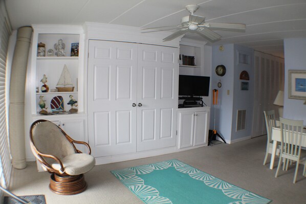 A room with a white wardrobe, a wicker chair, a teal rug, a ceiling fan, a television, and a dining table with chairs. - Studio & 1 Bath in Rehoboth Beach, DE -Ocean Block (Rehoboth Beach)