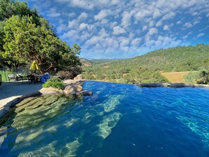 A clear blue pool with rock formations, surrounded by greenery and a view of distant hills. - Welcome to Sunset Hills Ranch! (Sutter Creek)
