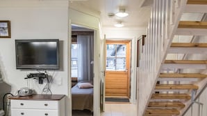 A room with a television, a desk, a staircase, and a bed in the adjacent room.