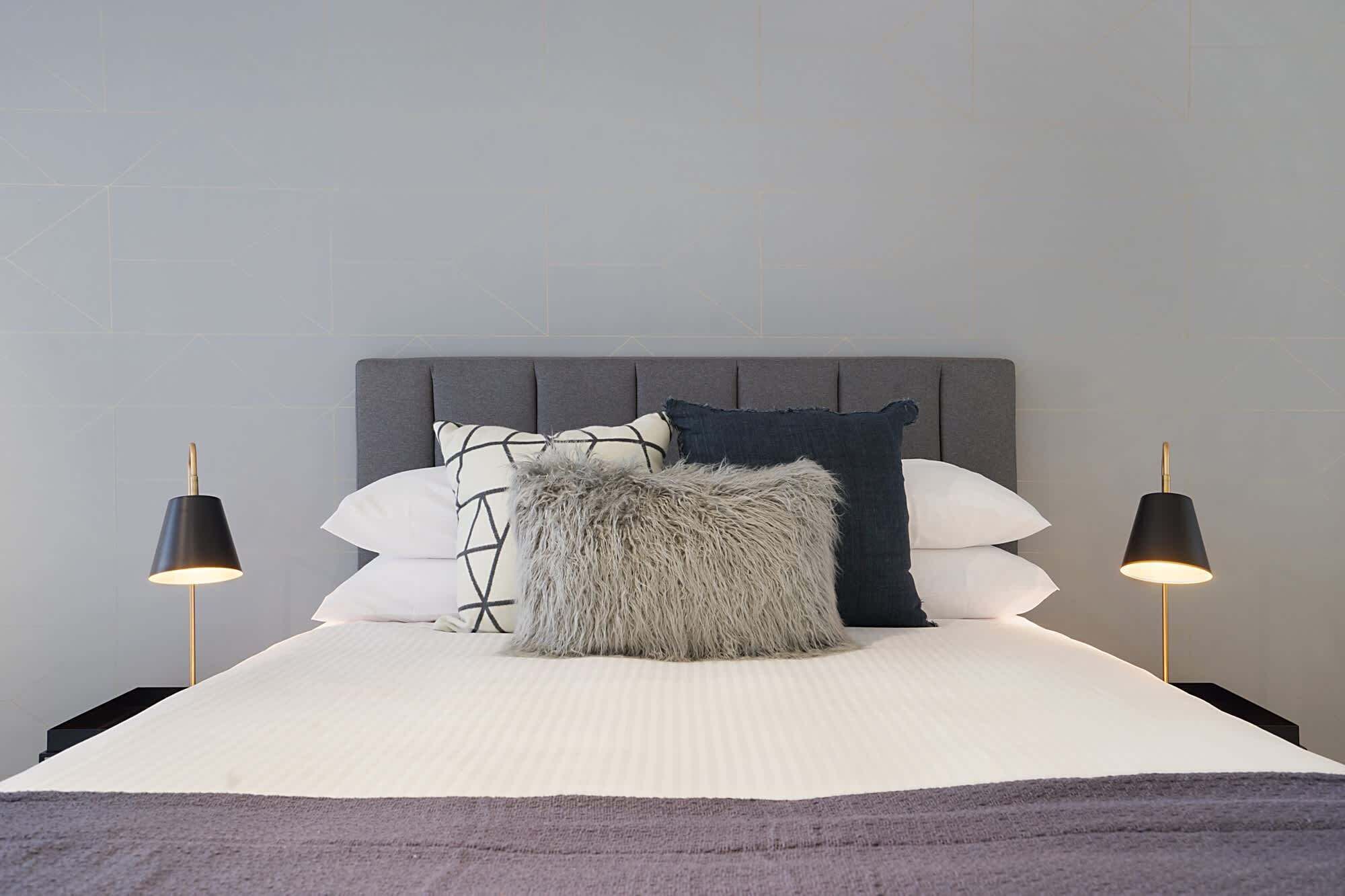 A neatly made bed with a grey headboard, white and patterned pillows, and a fluffy grey throw. Two black lamps are on either side of the bed.