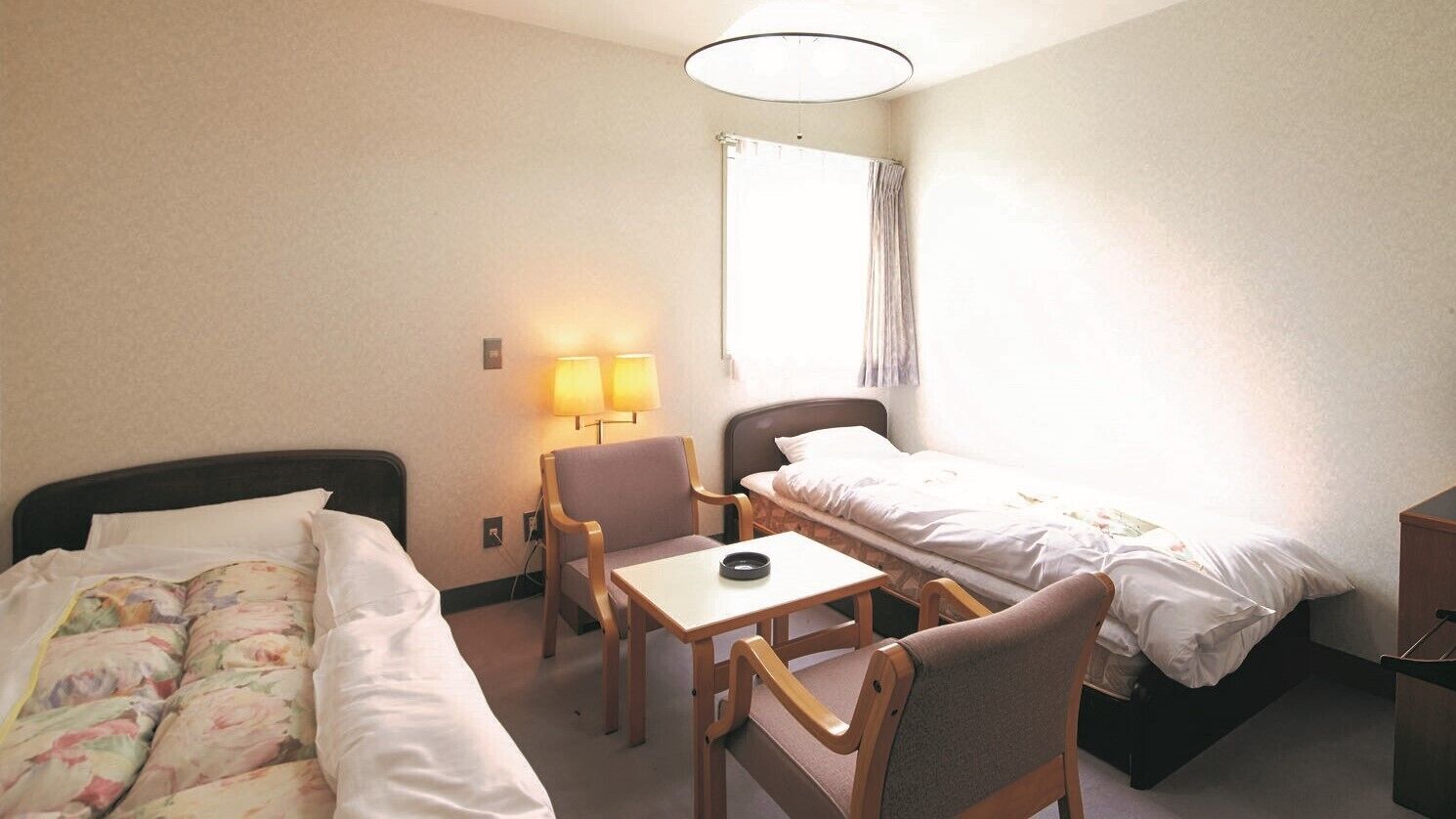 A hotel room with two beds, a small table, a chair, a lamp, and a window with curtains.