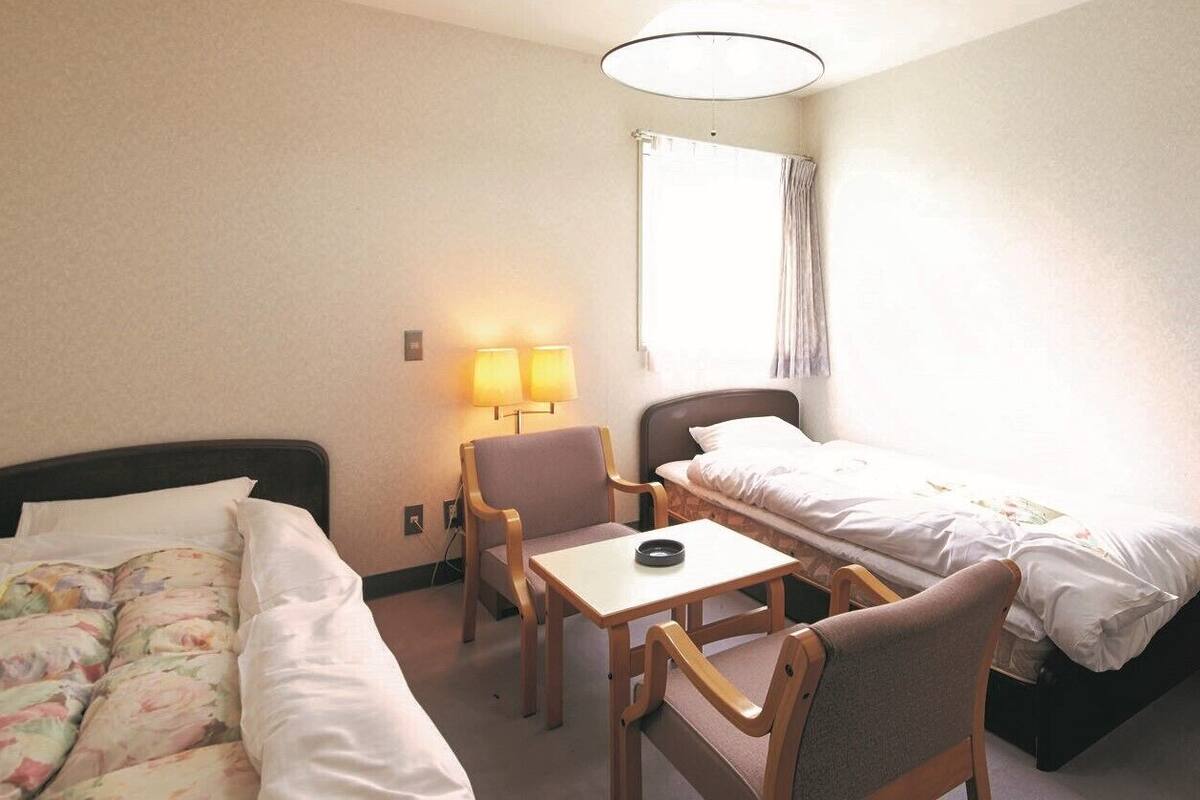 Une chambre d’hôtel avec deux lits, une petite table, une chaise, une lampe et une fenêtre avec des rideaux.