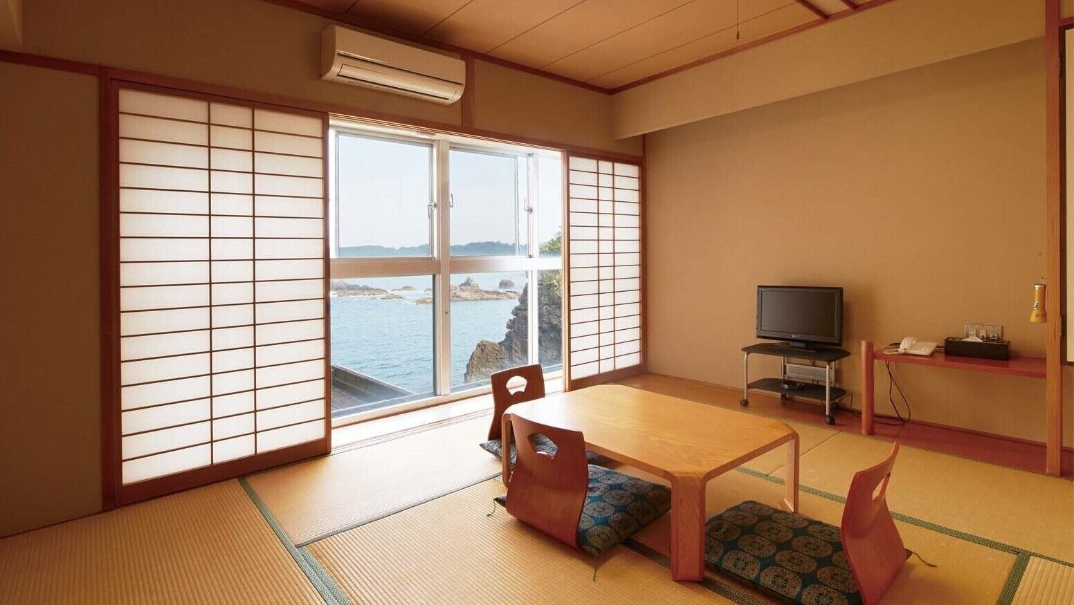 A traditional Japanese room with tatami flooring, a sliding door with a view of the sea, a television, and a small table.