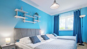 A bedroom with two beds, a nightstand, a lamp, a ceiling fan, and a window with curtains. - Nostalgia by Interhome (Mielno)