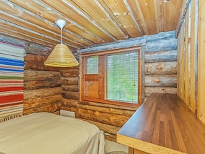 A log cabin room with a bed, a hanging light fixture, a window with blinds, and a wooden table.