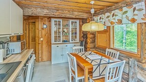 A rustic kitchen with wooden walls, a dining table with chairs, a cabinet with glass doors, and a window with a view of trees.