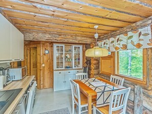 A rustic cabin interior with a wooden dining table, white chairs, a kitchen area with cabinets, and a window with a leaf-patterned curtain.