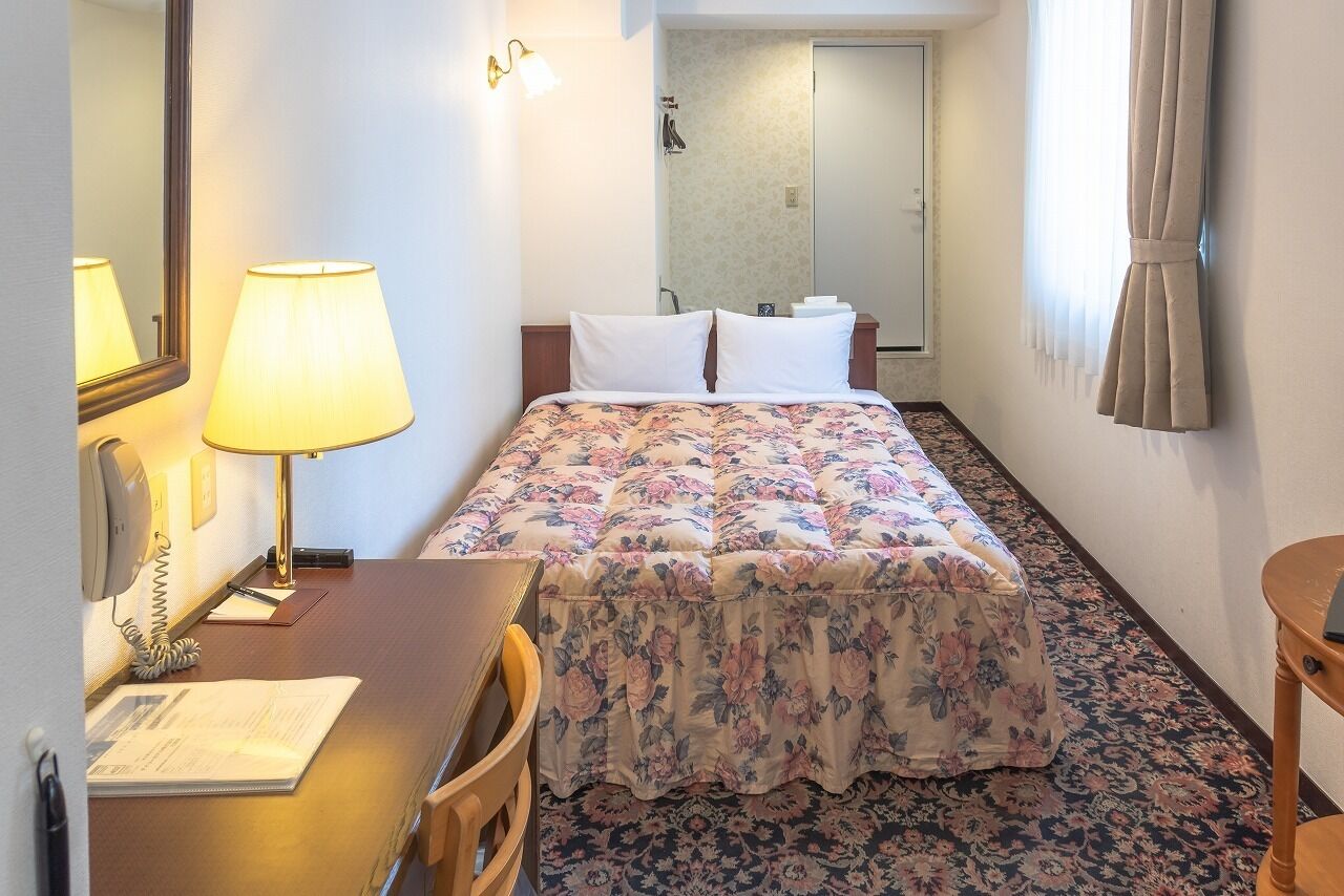 A hotel room with a bed, a desk with a lamp, a telephone, and a mirror.