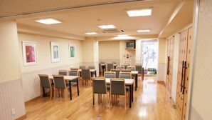 A spacious dining area with wooden tables and chairs, a long corridor with lockers, and framed pictures on the walls. - Asaka Daily Hotel (Asaka)