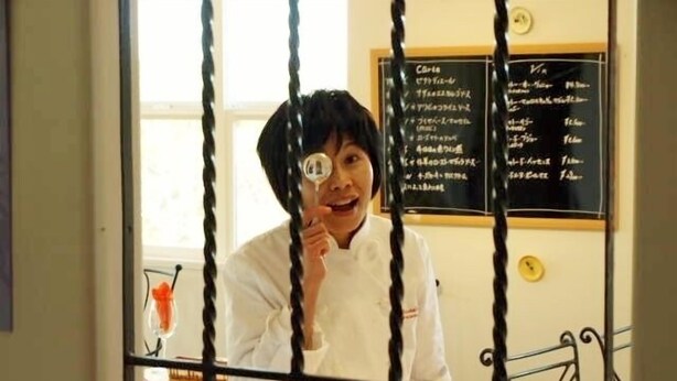 A person in a white chef's coat standing in front of a blackboard with writing, a bed with a yellow cover, and a window in the background. - Hostellerie Avances Soiree (Tateyama)