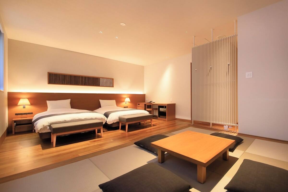 A modern hotel room with two beds, a small desk, a wooden table, and a sliding door.