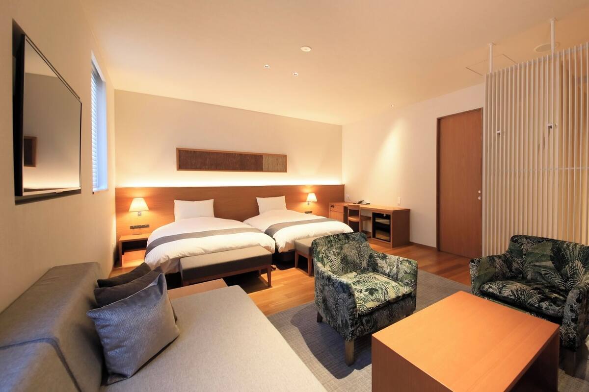A hotel room with two beds, a sofa, two armchairs, a desk, and a sliding door.