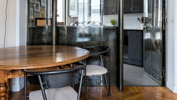 A dining area with a wooden table and chairs, a lamp, and a view into the kitchen through a glass partition.