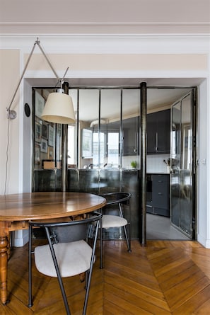 A dining area with a wooden table and chairs, a lamp, and a view into the kitchen through a glass partition.