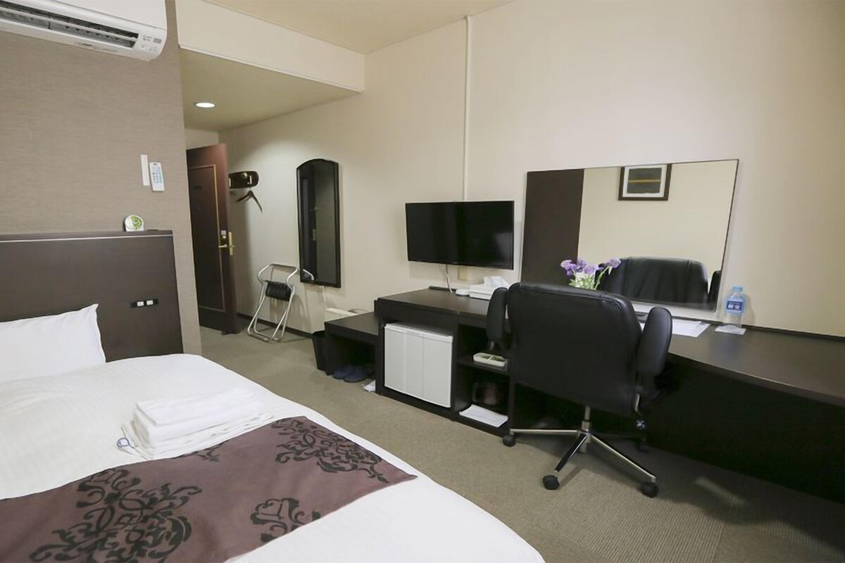 A hotel room with a large bed, a desk with a computer, a chair, a TV, and a mirror.