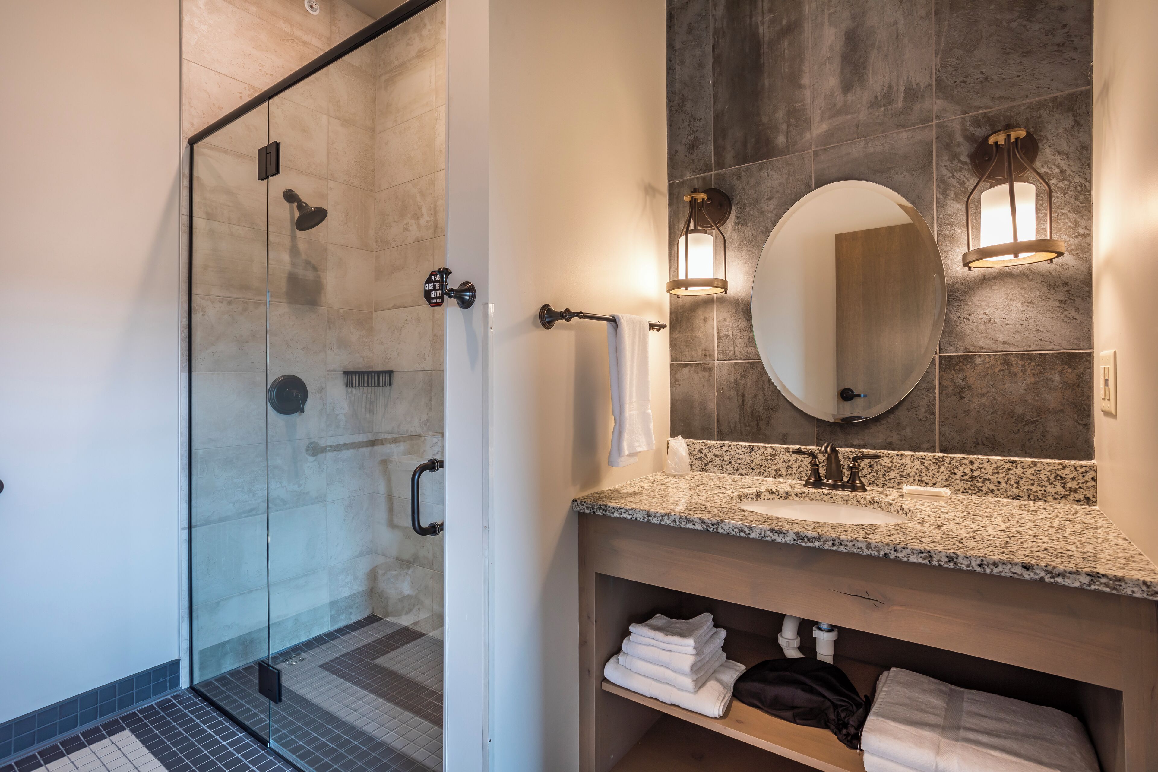 A modern bathroom with a glass-enclosed shower, a granite countertop sink, and a large mirror with wall-mounted lights.