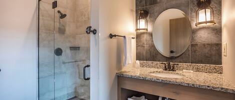 A modern bathroom with a glass-enclosed shower, a granite countertop sink, and a large mirror with wall-mounted lights.
