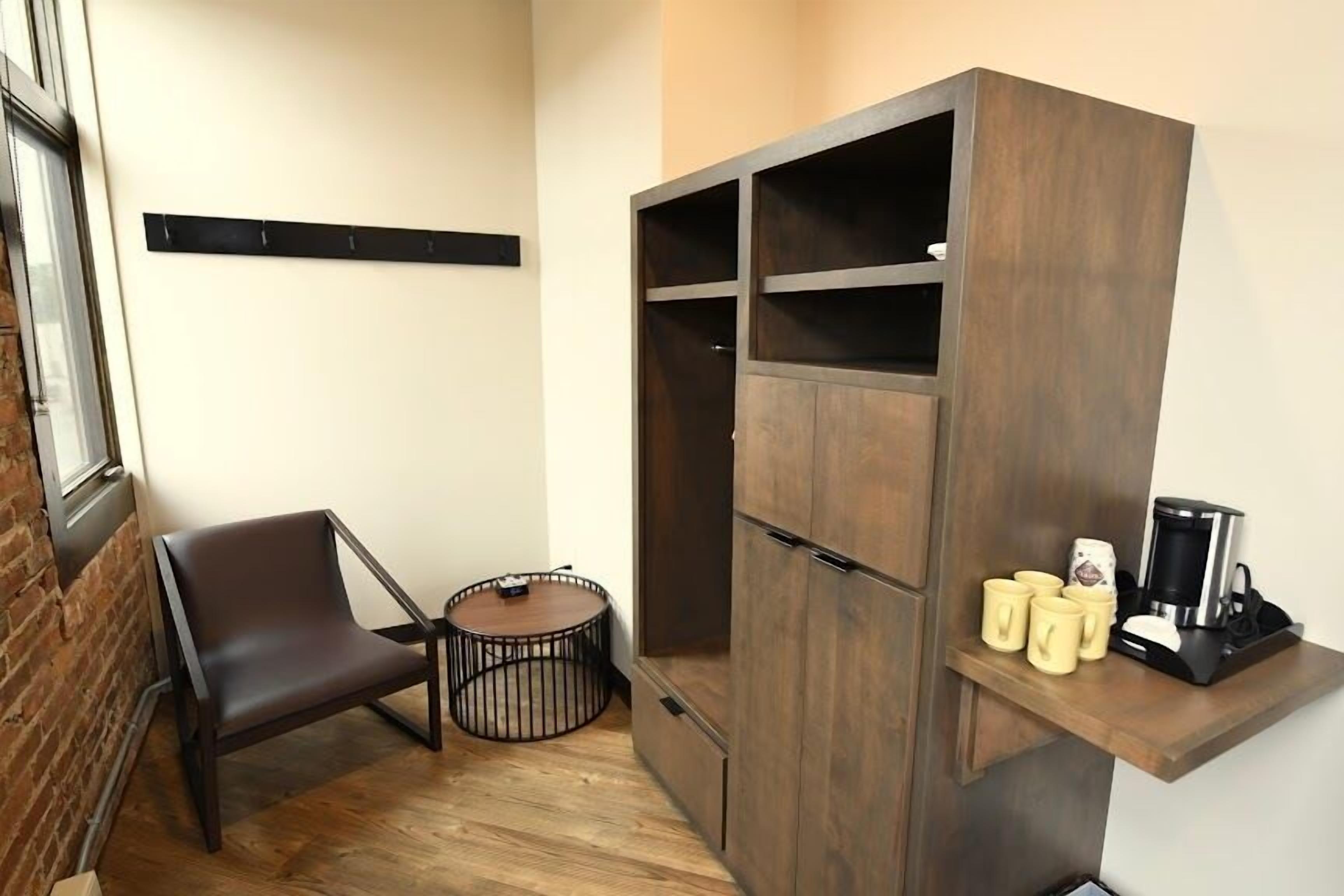 A small room with a dark brown wardrobe, a single chair, a round table, and a coffee maker on a shelf.