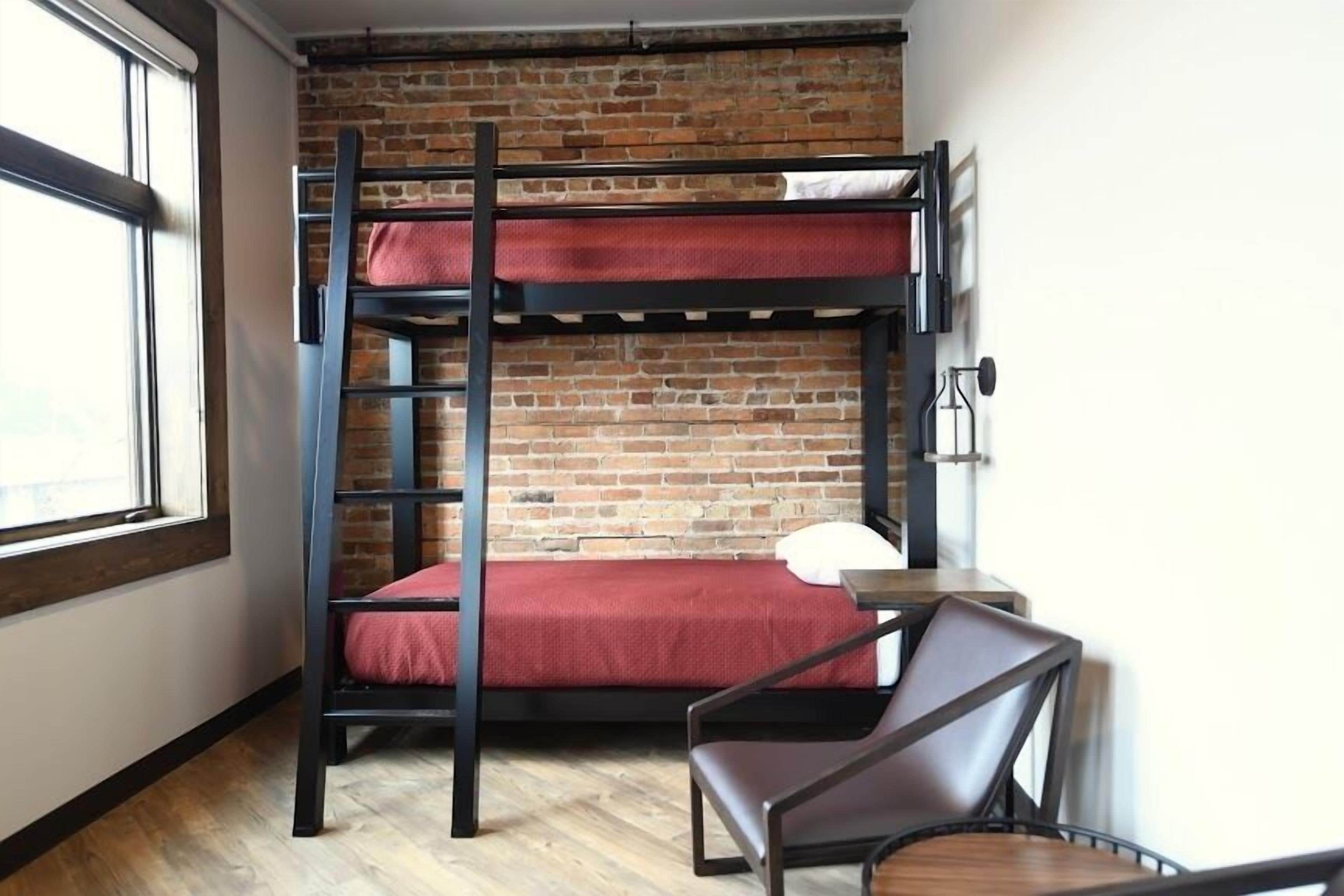 A bunk bed room with a brick wall, a wooden floor, a desk with a chair, and a window.