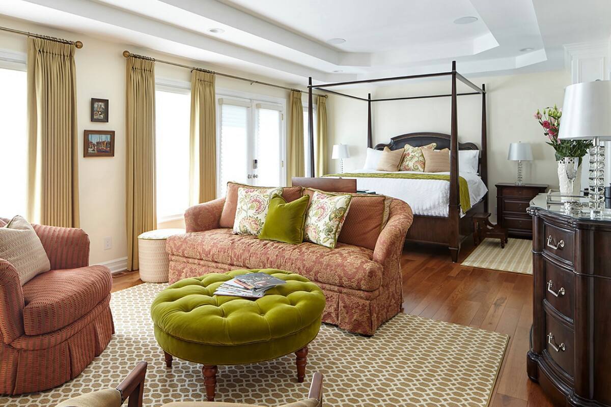 A spacious bedroom with a four-poster bed, a sitting area with a sofa and ottoman, and a dresser with drawers.