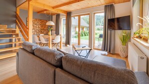 A spacious living room with a wooden ceiling, a gray sofa, a dining area with a table and chairs, and a large TV on the wall. - Chaty Pod Beskidkiem by Interhome (Szczyrk)