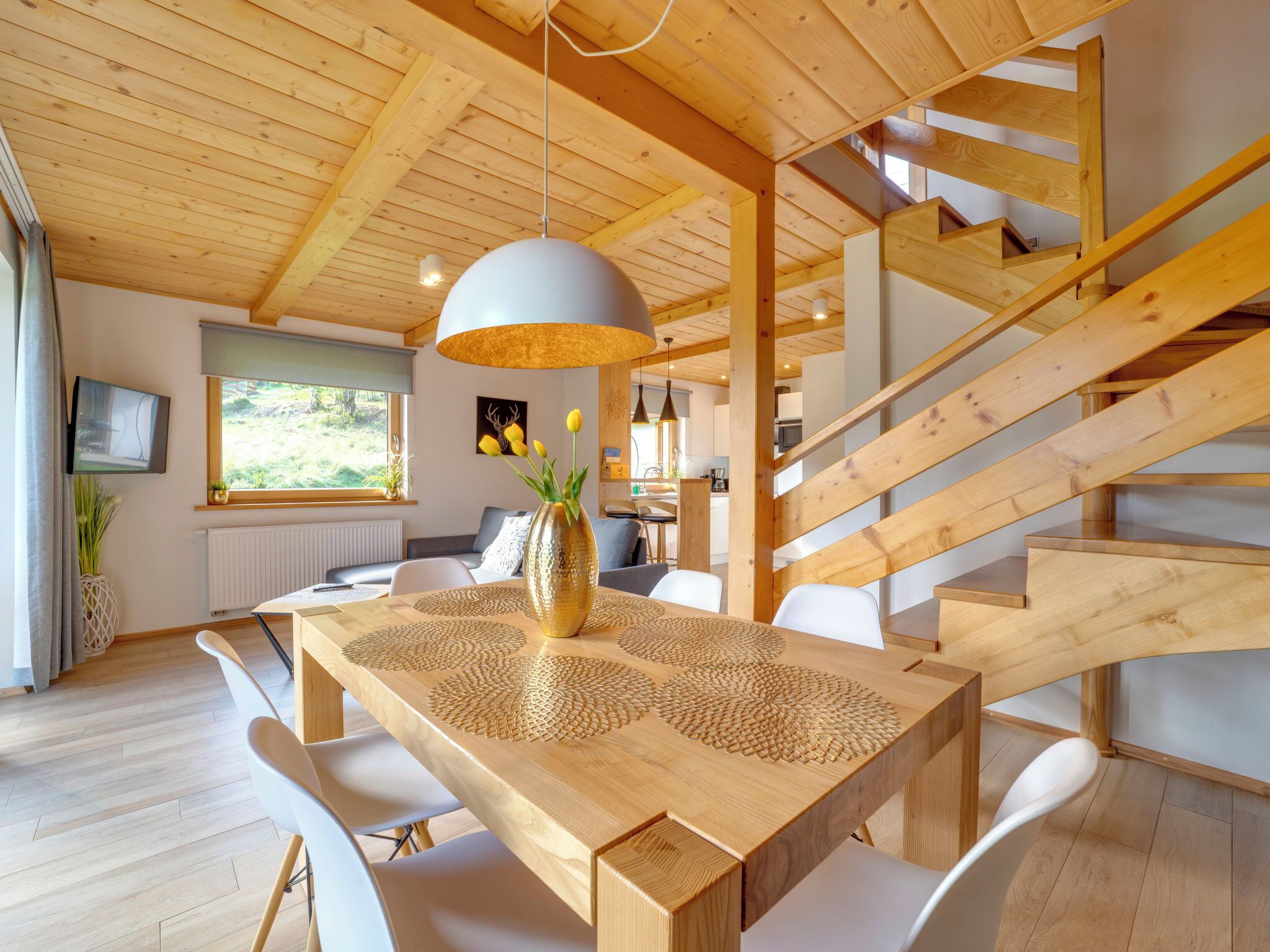 Un intérieur moderne avec une table à manger en bois, des chaises blanches, un escalier et une grande fenêtre.