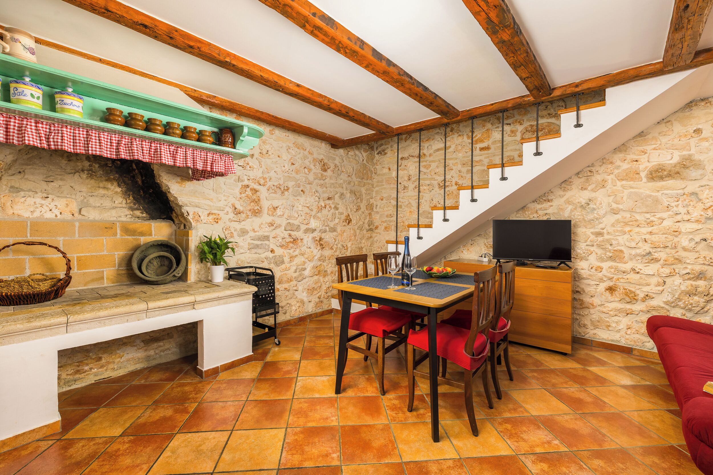 Una cocina rústica con pared de piedra, vigas de madera, una mesa de comedor con sillas y un televisor sobre un mueble de madera.