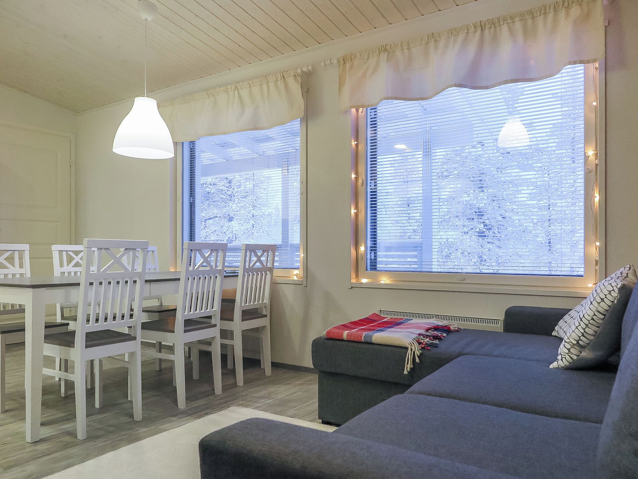 A room with a dining table, chairs, a sofa, and large windows with blinds.