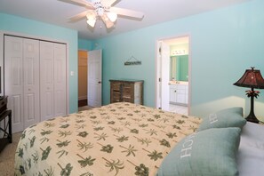A bedroom with a bed, a ceiling fan, a nightstand, a lamp, and a door leading to a bathroom. - Home with golf cart,community pool,Spinnakers Reach community (Emerald Isle)
