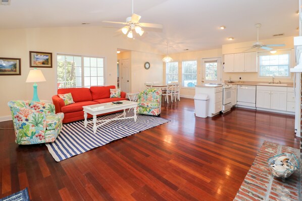A spacious living room with a red sofa, a white coffee table, and a striped rug. There is a kitchen area with white cabinets and a dining table. - Home with golf cart,community pool,Spinnakers Reach community (Emerald Isle)