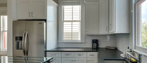 A modern kitchen with white cabinets, a stainless steel refrigerator, and a window with blinds.