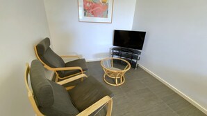 Ein Wohnzimmer mit zwei Holzstühlen, einem Korbstuhl, einem Flachbildfernseher auf einem Podest und einem Bilderrahmen mit einem abstrakten Gemälde an der Wand.