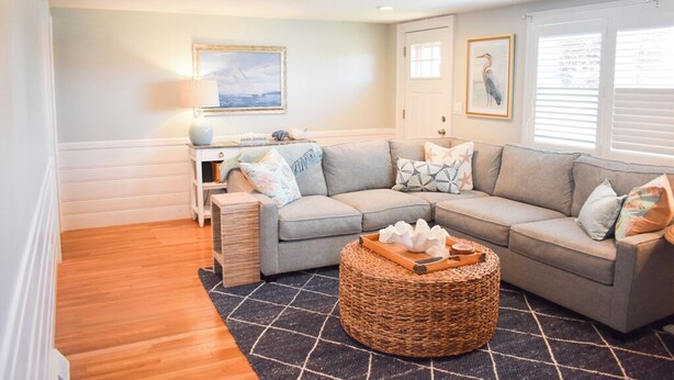 A living room with a grey sectional sofa, a round wicker coffee table, and a black and white rug. There is a desk with a lamp and a framed picture on the wall.