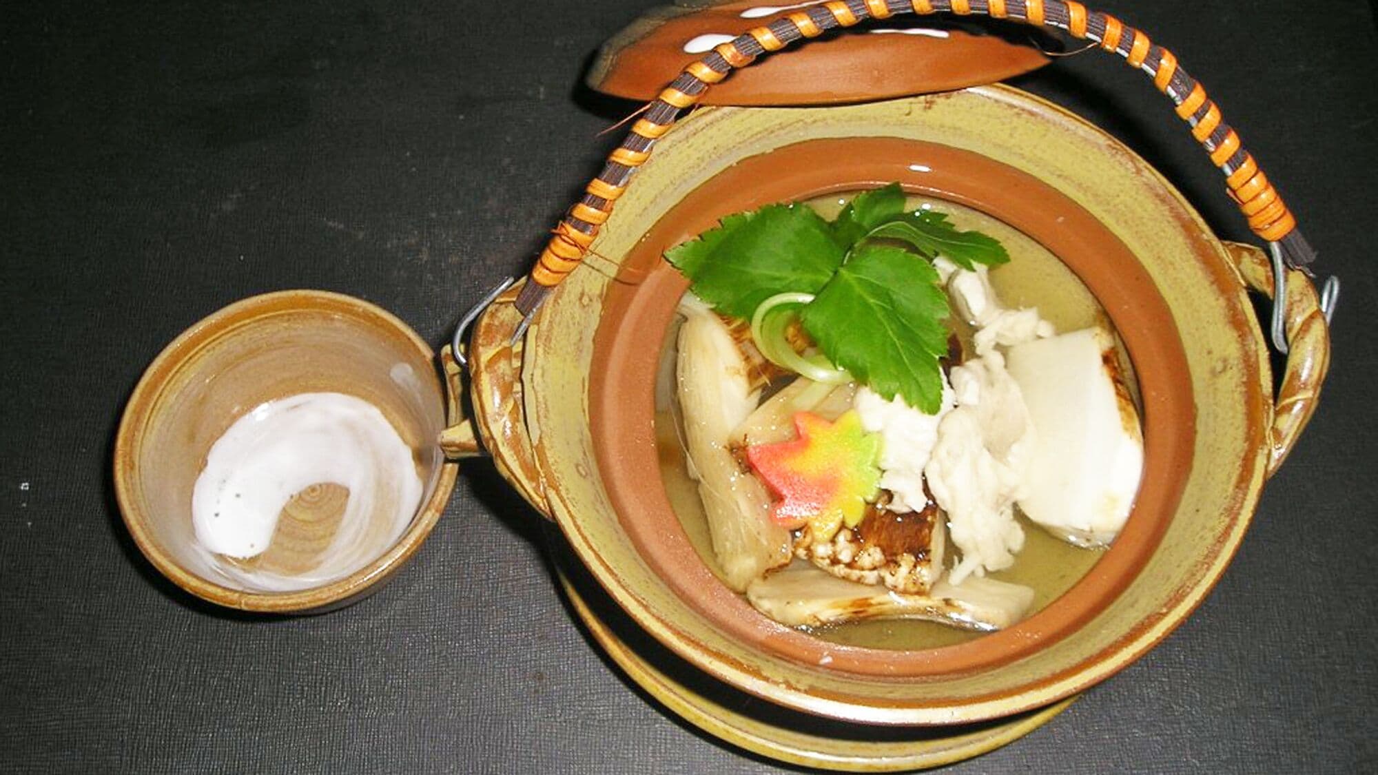 Een keramieken kom met handvat, gevuld met een gerecht, een groene bladgarnituur, en een kleine kom met een witte, romige substantie.