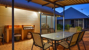 An outdoor patio with a grill, seating, and a view of a house with a blue roof. - Osprey Holiday Village Unit 117 - Villa with a Pool in the Complex (EXMOUTH)