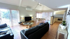 A spacious living area with a black sofa, a glass coffee table, a TV, and a dining area with chairs and a table. - Osprey Holiday Village Unit 117 - Villa with a Pool in the Complex (EXMOUTH)