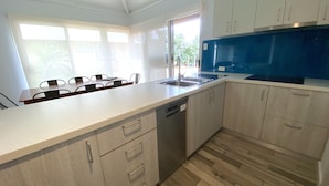 A modern kitchen with a dishwasher, a sink, and a blue microwave. There is a dining area with chairs and a table visible through the window. - Osprey Holiday Village Unit 121 - Villa with a Pool in the Complex (EXMOUTH)