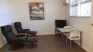 A room with a black leather sofa, a small table with a chair, a TV, and a painting on the wall.
