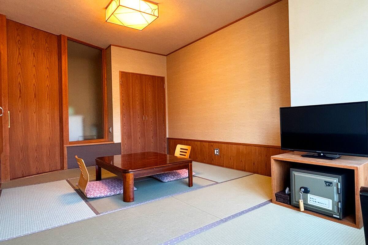 A room with a wooden wardrobe, a television on a wooden stand, tatami mats on the floor, and a small cabinet with a television.