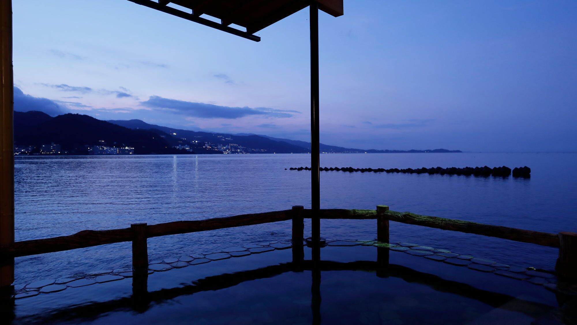 Vista al agua con muelle de madera, un horizonte urbano distante y montañas al fondo.
