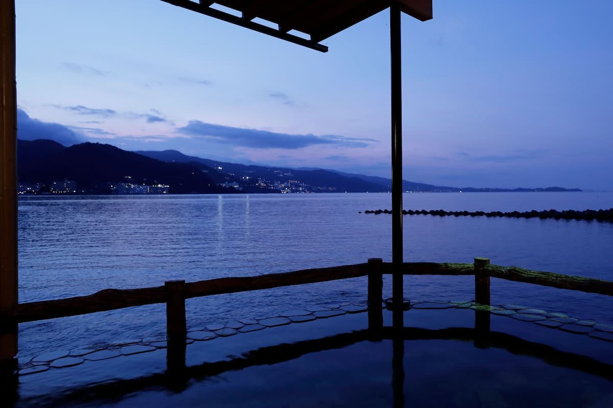 Vista al agua con muelle de madera, un horizonte urbano distante y montañas al fondo.