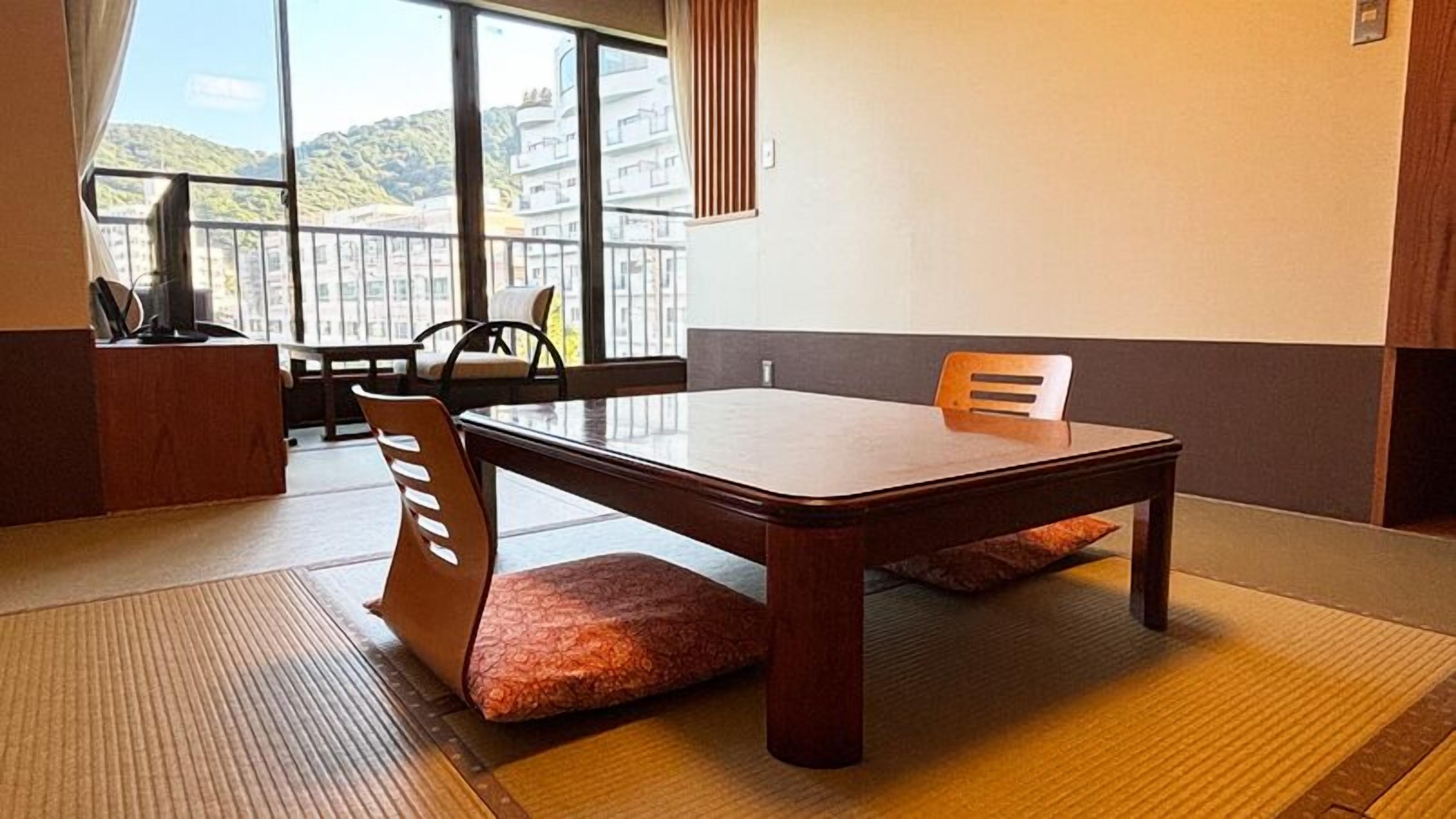 Una habitación con esteras tatami, una mesa baja, sillas, vista a edificios a través de ventanas grandes y un armario de madera.