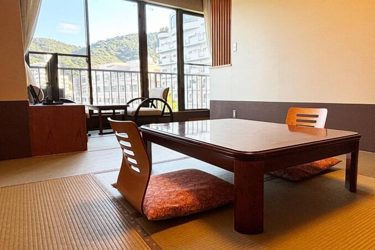 A tatami mat room with a low table and chairs, a view of buildings through large windows, and a wooden cabinet.
