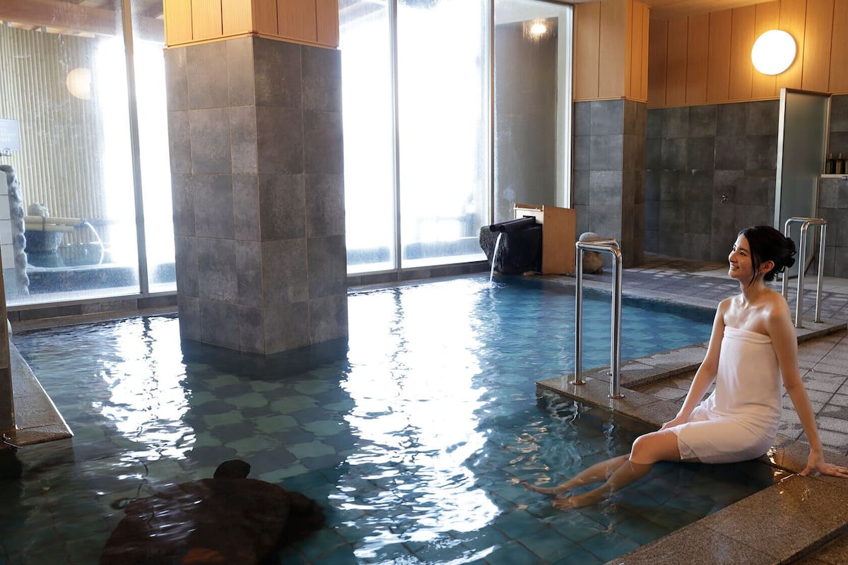 A person sitting by a pool with a stone wall and a window in the background.