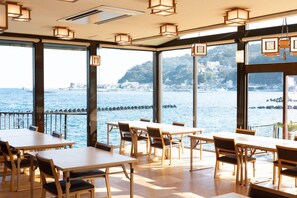 A restaurant with large windows offering a view of the sea and a hilly landscape. Tables and chairs are arranged for dining. - Atami Onsen Hiratsuru (Atami)