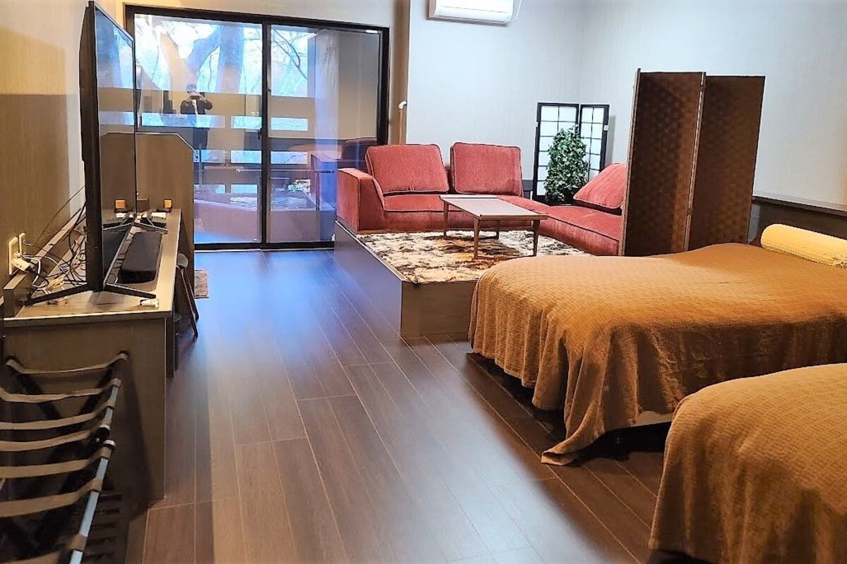A hotel room with a desk, two beds, a sofa, and a sliding glass door to a balcony.
