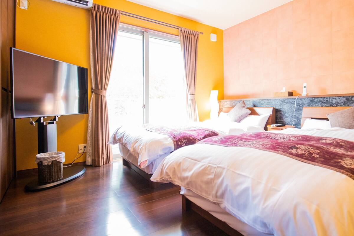 Una camera d'albergo con due letti, una televisione a schermo piatto, un cestino e una finestra con tende.