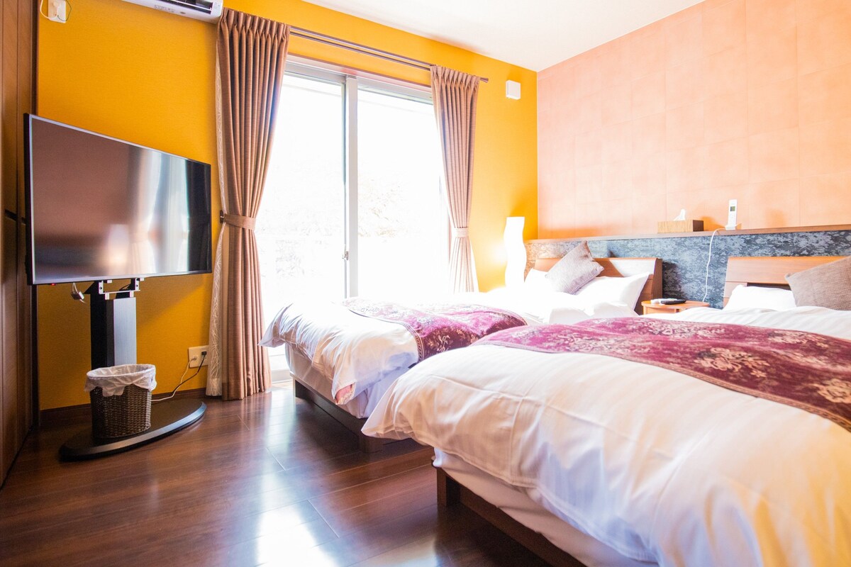 A hotel room with two beds, a flat-screen TV, a waste basket, and a window with curtains.