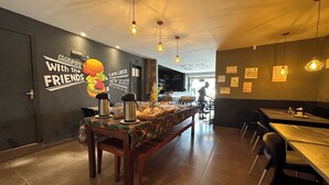 A restaurant interior with a long communal table, chairs, hanging pendant lights, and a wall with a mural and text. - Hotel Xenios (Macapa)
