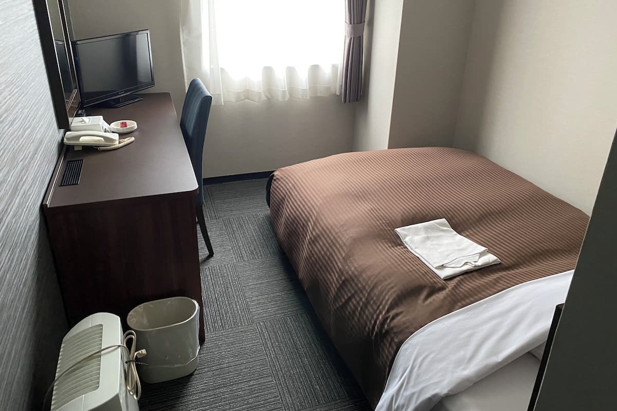 A hotel room with a bed, a desk with a telephone, a television, and a waste bin.